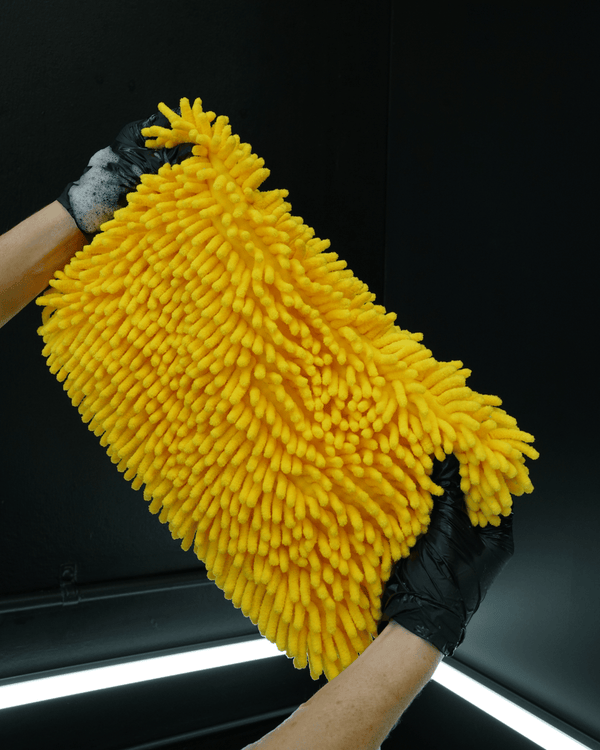 A detailer wearing black gloves holding a large noodle car wash mitt on a black background