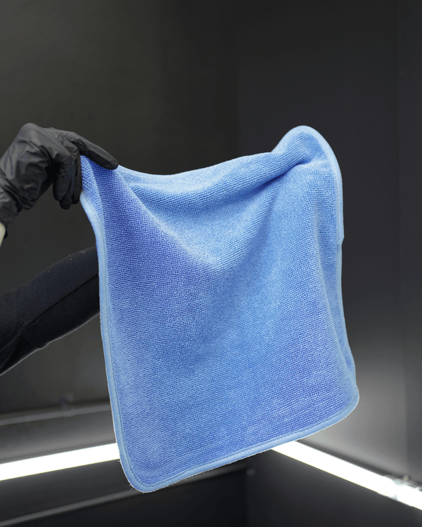 A person wearing black gloves holding a blue microfibre drying towel on a dark background
