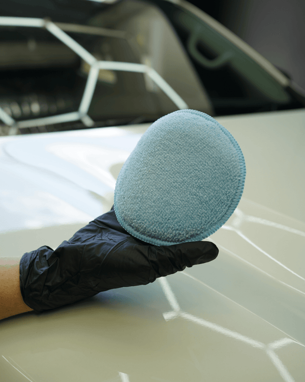 A person with black gloves on holding a polish and wax applicator on a white car bonnet in a detailing bay