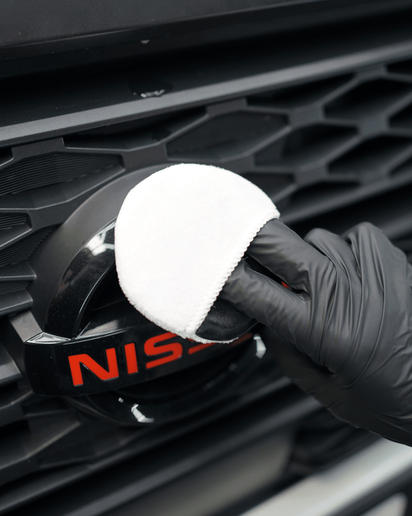 Person cleaning a Nissan car emblem with a white applicator and black glove.