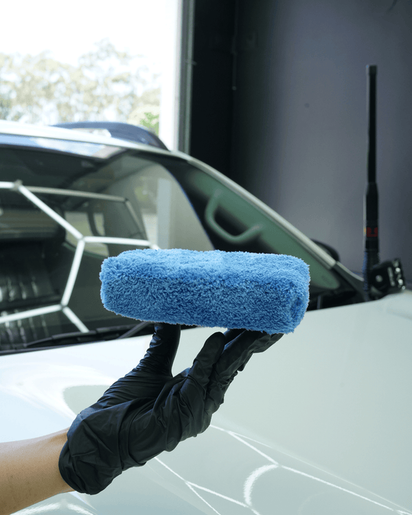 A person wearing a black glove holding a plush applicator sponge next to a white car in a detailing bay