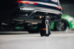 A black hand pump pressure sprayer on the ground with a black and green car in the background