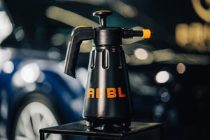 A black pressure foam cannon on a stool with a car in the background 