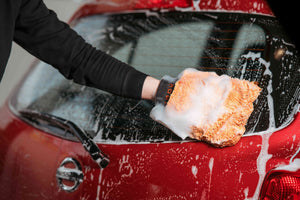 A person using a soapy orange microfibre wash mitt to wash a red car
