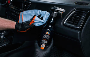 A person wearing black gloves holding a blue microfibre cloth spraying a car dashboard