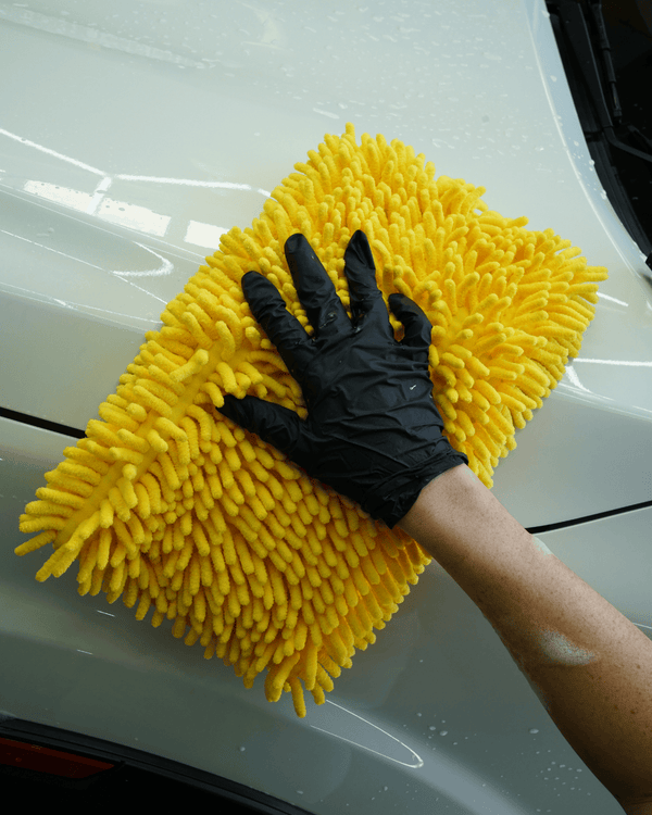 A detailer wearing black gloves washing a white car with a large yellow noodle sponge 