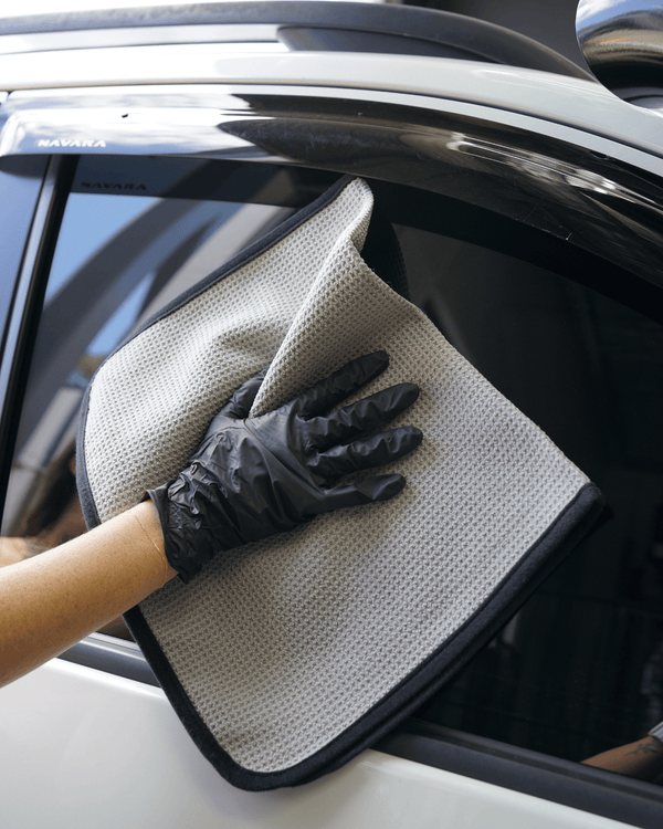 A person with black gloves on cleaning a car window with a grey waffle weave towel