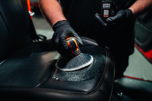 Hand using ADBL Fat Brush to clean and agitate foam cleanser on a black leather car seat and upholstery