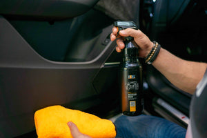 Hand holding ADBL INTERIOR WOW protectant bottle and orange microfibre cloth next to car door panel