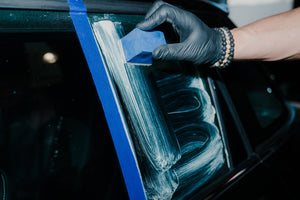 Close-up of a hand polishing car glass with ADBL Glass Polish and a blue applicator, taped area visible