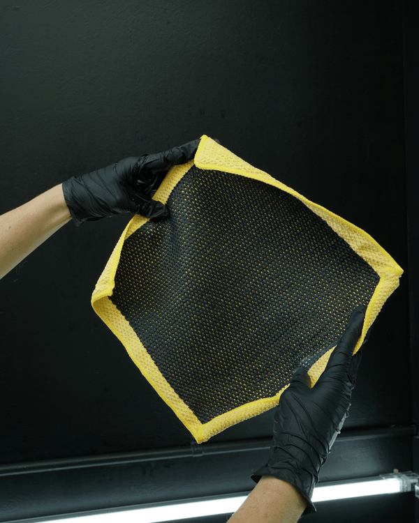 A detailer wearing black gloves holding a yellow clay cloth on a black background 