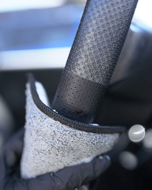 A person with a black glove on holding a car interior scrub sponge cleaning a steering wheel