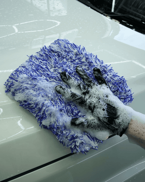 A detailer wearing black gloves washing a white car bonnet with a large blue microfibre wash pad 