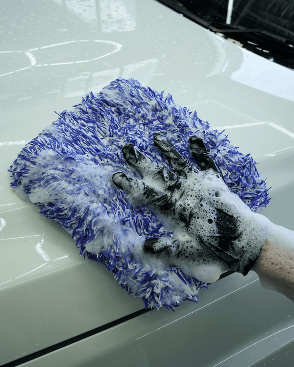 A detailer wearing black gloves washing a white car bonnet with a large blue microfibre wash pad 