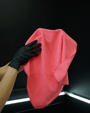 Person wearing a black glove holding a pink microfibre cloth against a dark background