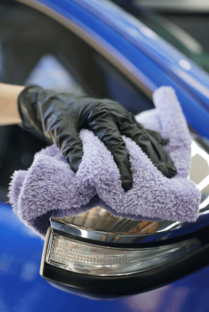 Person cleaning a blue car mirror with a purple microfibre cloth and black gloves.