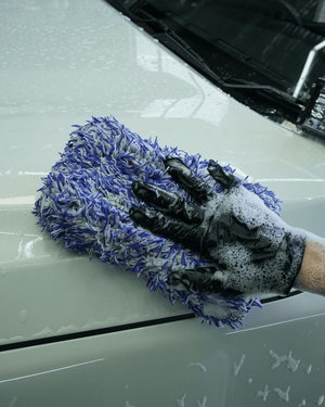 A detailer wearing black gloves washing a white car bonnet with a blue and white microfibre wash pad.