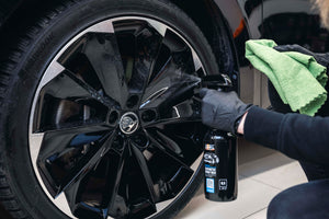 Detailer cleaning a glossy black car wheel with ADBL Synthetic Spray Wax and a green microfibre cloth for added protection.