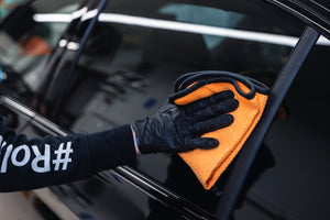 Hand in glove wiping black car interior window trim with orange microfibre cloth