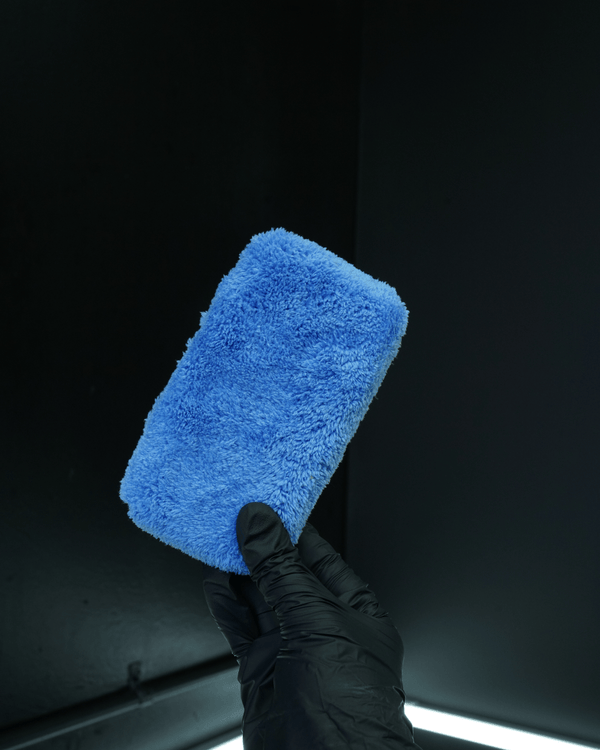 Blue microfibre applicator held by a gloved hand against a dark background