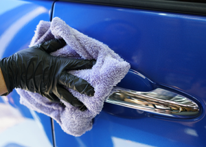 Person cleaning a blue car door handle with a purple microfibre cloth and black gloves.