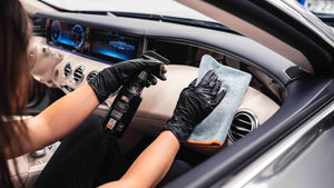 Person applying ADBL INTERIOR QD with a spray bottle and grey and orange microfibre towel to a car dashboard